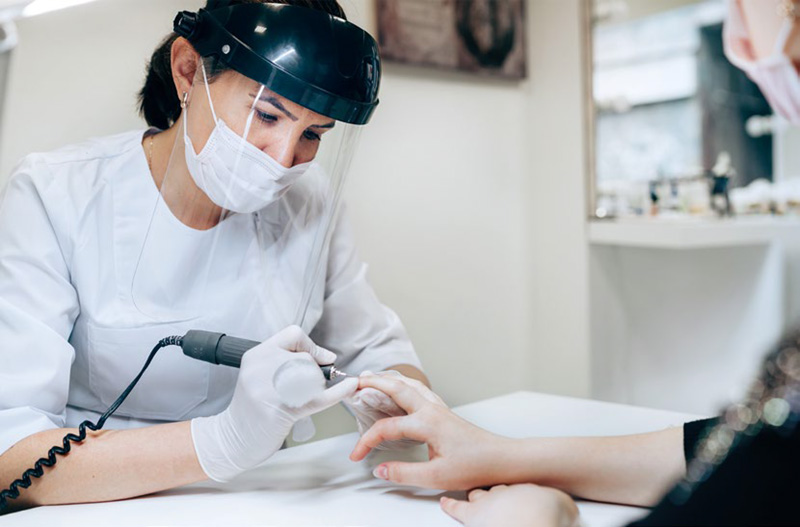 a nail technician wears a mask and face shield for protection while she works on a client's hand