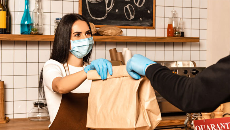 a barista wearing a face mask and rubber gloves hands a brown bag to a customer also wearing gloves