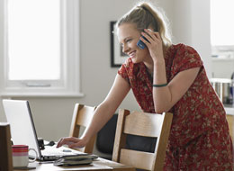 woman leaning over chair working on laptop while also talking on phone