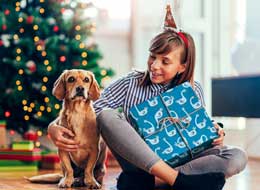 girl holding a gift and hugging a dog in front of a christmas tree